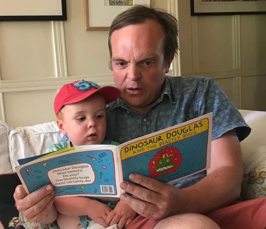 A father reading Dinosaur Douglas and the Beastly Bugs with a toddler in a red cap