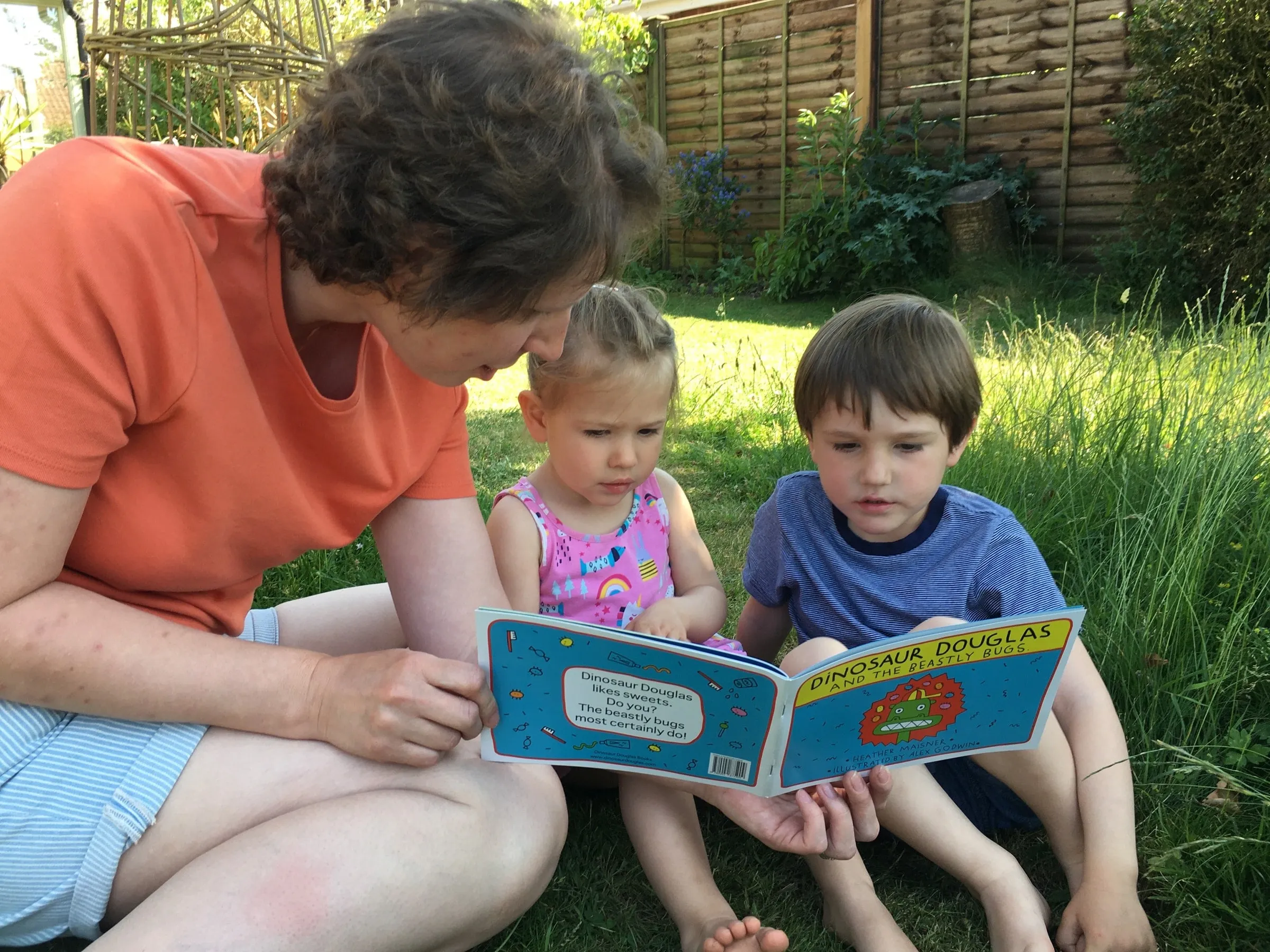 A mother reading Dinosaur Douglas and the Beastly Bugs to two young children on the grass